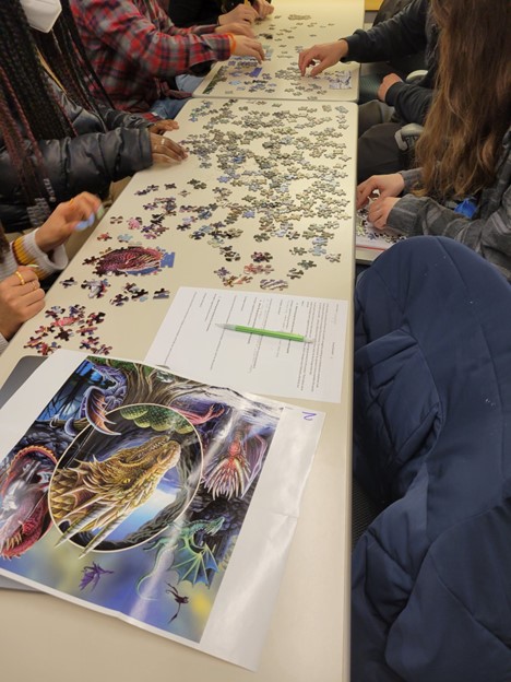 A photo of students assembling puzzle pieces. They have a piece of paper showing the completed puzzle image. It is an illustration of dragons.